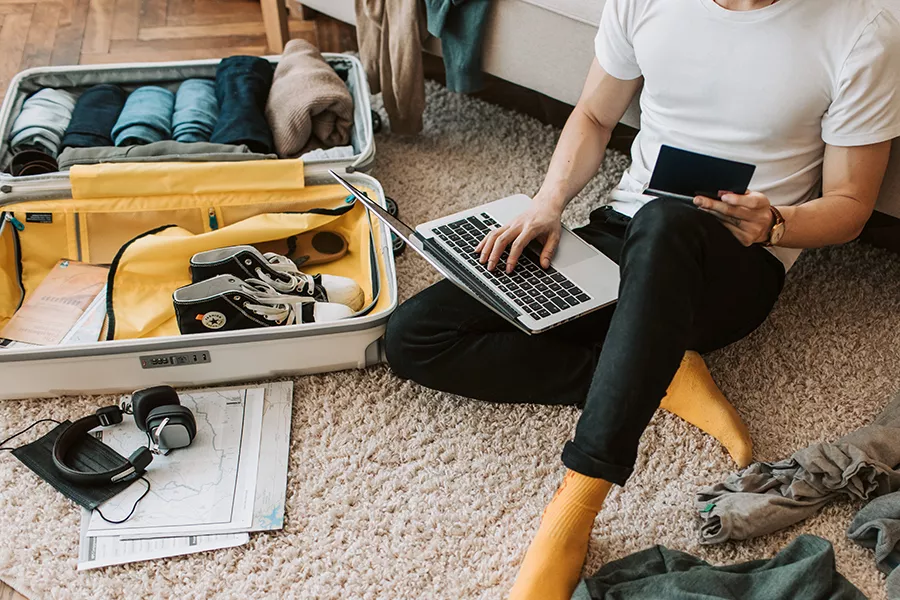 Person sitting on the floor with an open travel suitcase consulting travel information on their laptop